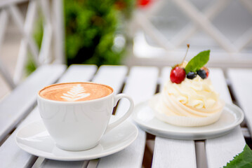 A cup of cappuccino and appetizing white meringue, decorated with fresh berries, on a white wooden table in a summer cafe.