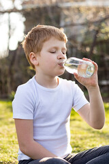 A red-haired teenager boy in a white T-shirt meditating in nature, doing yoga, resting and drinking water sitting on the grass at sunset in the park