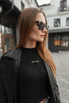 Stylish beautiful young woman with cool sunglasses wearing trendy black mockup outfit with a black leather coat jacket and T-shirt walks in the city