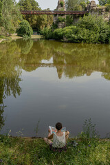 Paris, France - 06 18 2022: Park des Buttes Chaumont. A woman sitting from behind, reads a newspaper by the lake