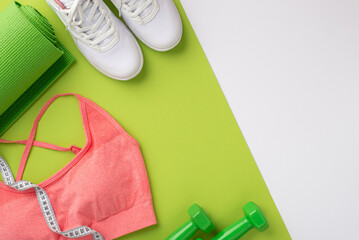 Fototapeta na wymiar Sports concept. Top view photo of white sneakers pink sports bra tape measure green exercise mat and dumbbells on bicolor green and white background with empty space