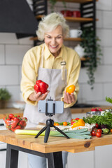 Senior woman cooking with video receipts or blogging. Mature female preparing fresh vitamin vegetarian salad with organic ingredients with a tutor in her smartphone. Elder using technology