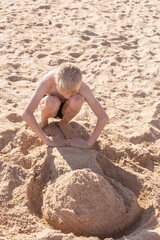 A fair-haired boy builds a sand tower on the beach on a summer sunny day. Happy holidays