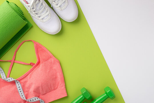Sports concept. Top view photo of white sneakers pink sports bra tape measure green exercise mat and dumbbells on bicolor green and white background with empty space