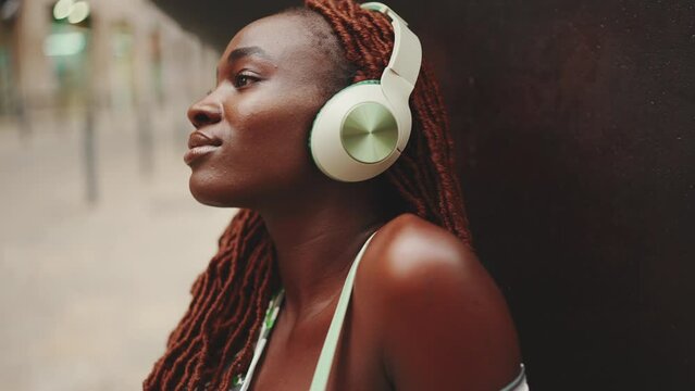 Close-up profile of beautiful woman with African braids in headphones enjoying music outside on the street. Stylish girl listening to music with her eyes closed