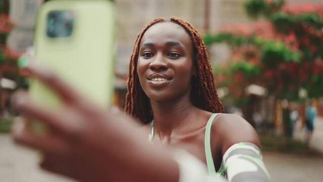 Smiling gorgeous woman with African braids wearing top stands outside on the street and uses mobile phone. Stylish girl taking selfie photos
