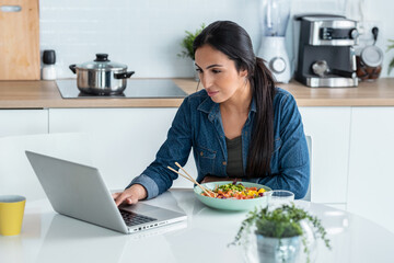 Cheerfull woman working with computer while with computer while eating poke bowl in the kitchen at...