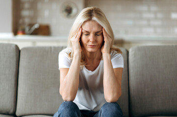 Middle aged blonde woman sits on couch at living room holding her head with her hands, feels...