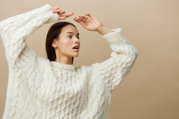 an elegant woman stands on a light background in a white sweater gently raises her arms above her head, creating rounded shapes and looks away with a slight dissociation, turning her body in the same