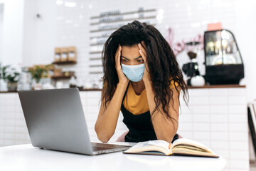 Upset African American small business owner, barista or waitress wearing medical mask sadly looks at laptop monitor, small business financial loss due to quarantine, bankruptcy