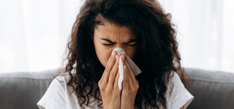 Unhealthy young woman. African american millennial girl is sick with flu, she sneezes and blows her nose in a napkin while sitting on the couch at home