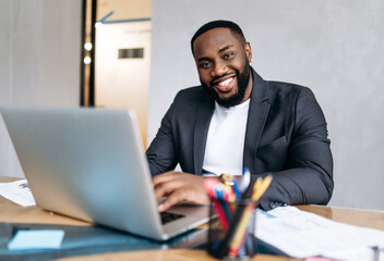 Successful male manager or boss is sitting at work desk in office. African american business man using laptop, develop new project or startup. Handsome confident employee look at the camera, smiling