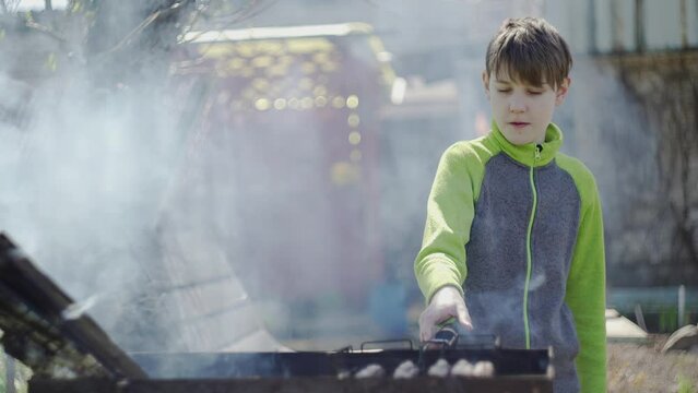 The boy is standing in front of the grill and turning over the sausages that are cooking on it. Shooting at a barbecue