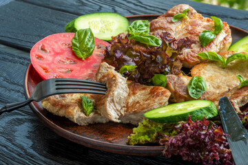 Pork chops. First piece is cut off. Close-up. In ceramic plate. Supplemented with basil leaves, lettuce, tomato and cucumber slices. On black background. Hot meat dish, Homemade
