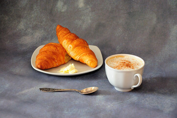 Two fresh croissants with butter on a plate and a cup of hot cappuccino with cinnamon on a gray background.