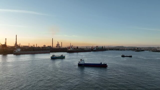 Small cargo ship sails by smokestacks to industrial harbor at sunset