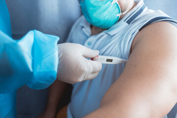 A nurse in PPE gear inserts a digital thermometer in the armpit of a man wearing a N95 mask....