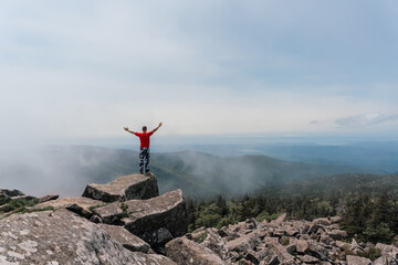 Hiker man on the top of the mountain enjoys the aerial view, raising his hands above the clouds. Mount Pidan