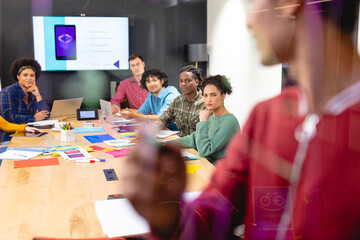 Young biracial male app developer explaining colleagues over chart on glass in board room