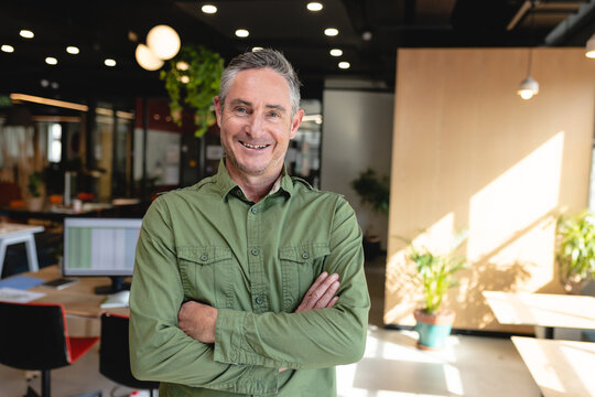 Portrait of smiling mature caucasian creative businessman standing with arms crossed in office