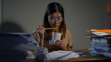 Asian businesswoman eating noodles cup junk food between working hard at the night looking the laptop computer technology in workplace at late overtime, work at home.