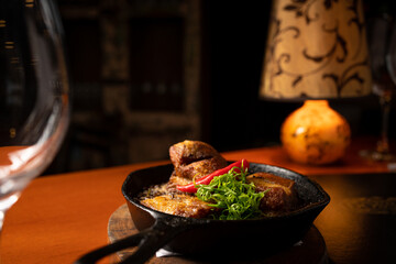 cast iron pan with stew, fried pork, lentil, onion and chili on wooden table with glasses, closed menu and warm light