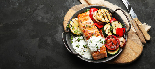Salmon steak with vegetables and white sauce on dark background.