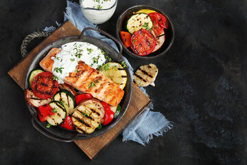Salmon steak with vegetables and white sauce on dark background.