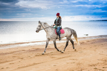 dressing jeans, jacket and spring hat lass hacks a dappled horse along seabeach