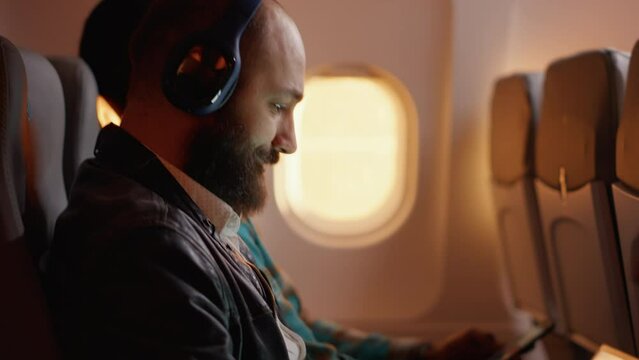 Tired employee travelling by airplane and working on laptop, using computer on flight during sunset. Work trip on plane, flying on vacation in economy class with people. Handheld shot.