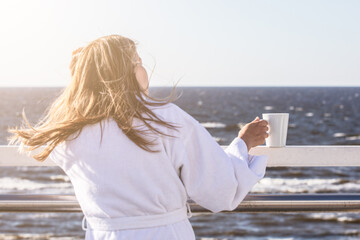 Fototapeta na wymiar A woman in a white robe with a white mug in her hands on the terrace looks at the sea, a romantic mood.