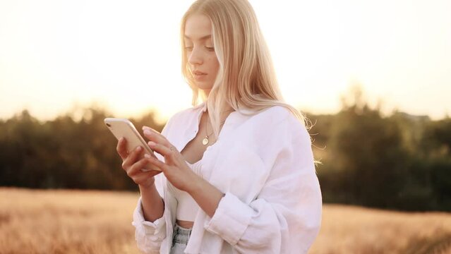 Young beatiful woman in white shirt texting on her phone in the gold field during the sunset.