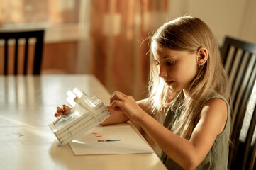 a 6-7-year-old girl with blonde hair in a green T-shirt is sitting at home at a table and drawing with markers. The girl is engaged in creativity. She's learning to draw
