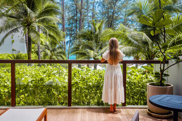Woman standing on balcony with tropical view. Back view of female traveler relax in modern hotel...
