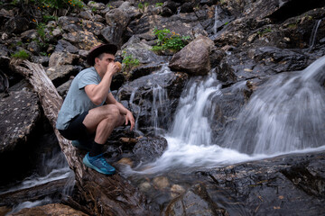 Young tourist in hat drinks from kuksa from waterfall in mountains