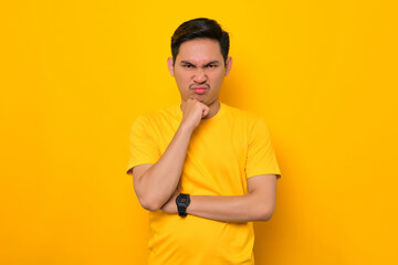 Annoyed young Asian man in casual t-shirt holding hand on chin, looking at camera isolated on yellow background. People lifestyle concept