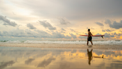 Healthy Asian senior man surfer in swimwear holding surfboard walking on the beach at summer sunset. Elderly retired male enjoy outdoor activity lifestyle and water sport surfing on summer vacation