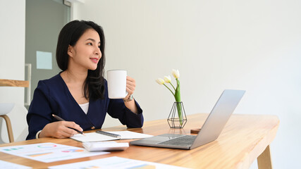 Attractive young business lady in stylish clothes holding a cup of coffee and looking out of office window