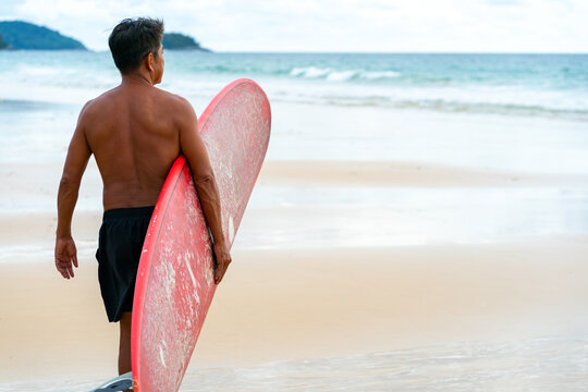 Healthy Asian senior man surfer in swimwear holding surfboard walking on the beach at summer sunset. Elderly retired male enjoy outdoor activity lifestyle and water sport surfing on summer vacation