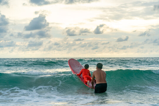Happy Asian family grandfather teaching grandchild boy surfing on surfboard in the sea on summer vacation. Senior man and little boy enjoy outdoor activity lifestyle and water sport surfing together.