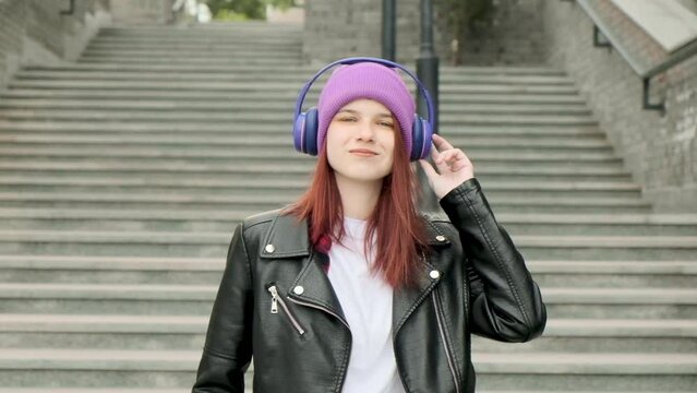 Caucasian happy beautiful girl wearing headphones, standing on street looking at camera and listening to music outdoors. joyful girl in good mood enjoys songs in city and laughs