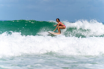 Asian woman surfer surfing and riding surfboard  the wave in the sea at tropical beach in sunny day. Healthy female enjoy outdoor activity lifestyle and water sport exercise surfing on summer vacation