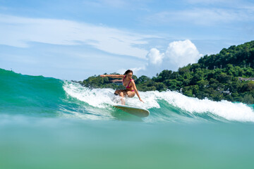 Asian woman surfer surfing and riding surfboard the wave in the sea at tropical beach in sunny day. Healthy female enjoy outdoor activity lifestyle and water sport exercise surfing on summer vacation