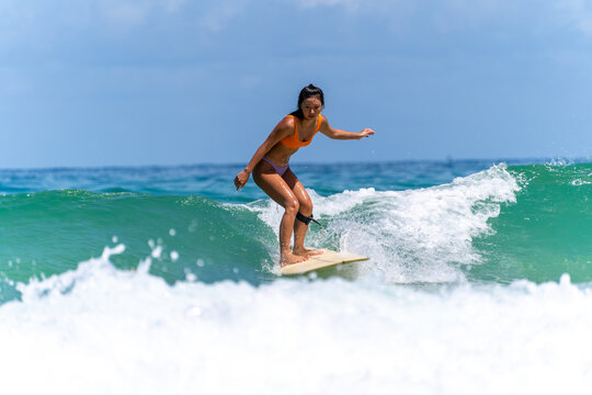 Asian woman surfer surfing and riding surfboard  the wave in the sea at tropical beach in sunny day. Healthy female enjoy outdoor activity lifestyle and water sport exercise surfing on summer vacation