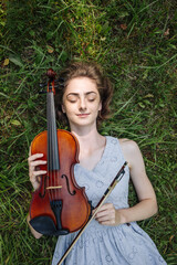 The girl lies with a violin on the grass in a city park.