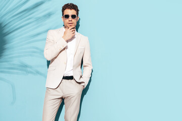 Portrait of handsome confident stylish hipster lambersexual model. Sexy modern man dressed in white elegant suit. Fashion male posing in studio near blue wall in sunglasses