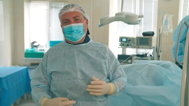 Mid-aged Male Doctor In Mask And Cap Equipped For The Surgery Talks To Camera. Surgeon Holding A Long Needle Instrument In His Gloved Hands.