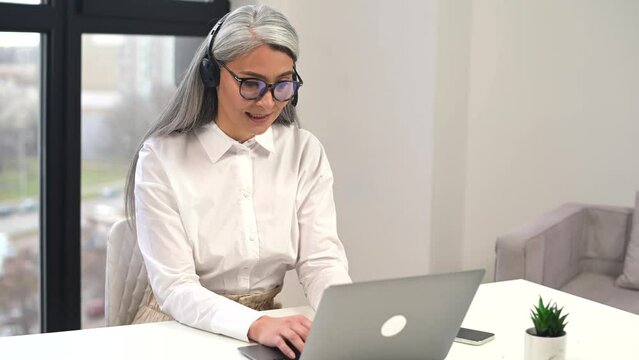 Confident aged woman with headset, takes a call, listening at the interlocutor, consulting the client online , gives the advice, working, positive person