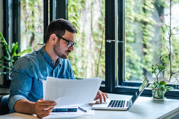 Serious businessman holding papers while scrolling business plan on laptop