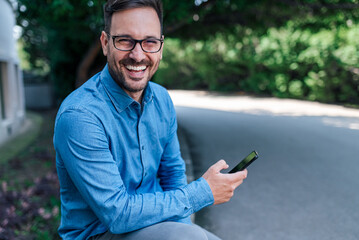 Portrait of cheerful businessman using smart phone while sitting by the road